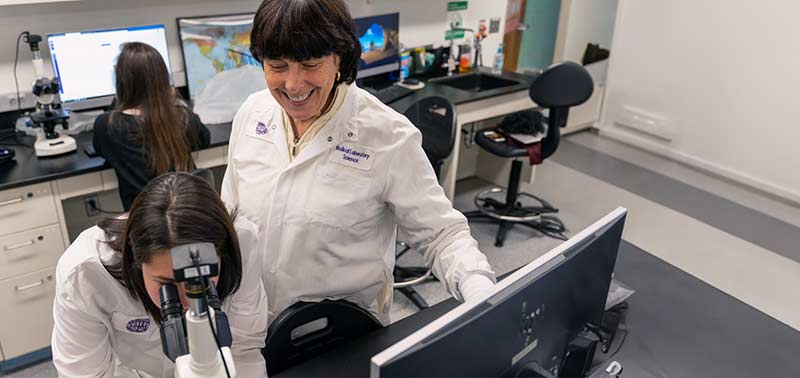 MLS professor smiling at student looking through a microscope